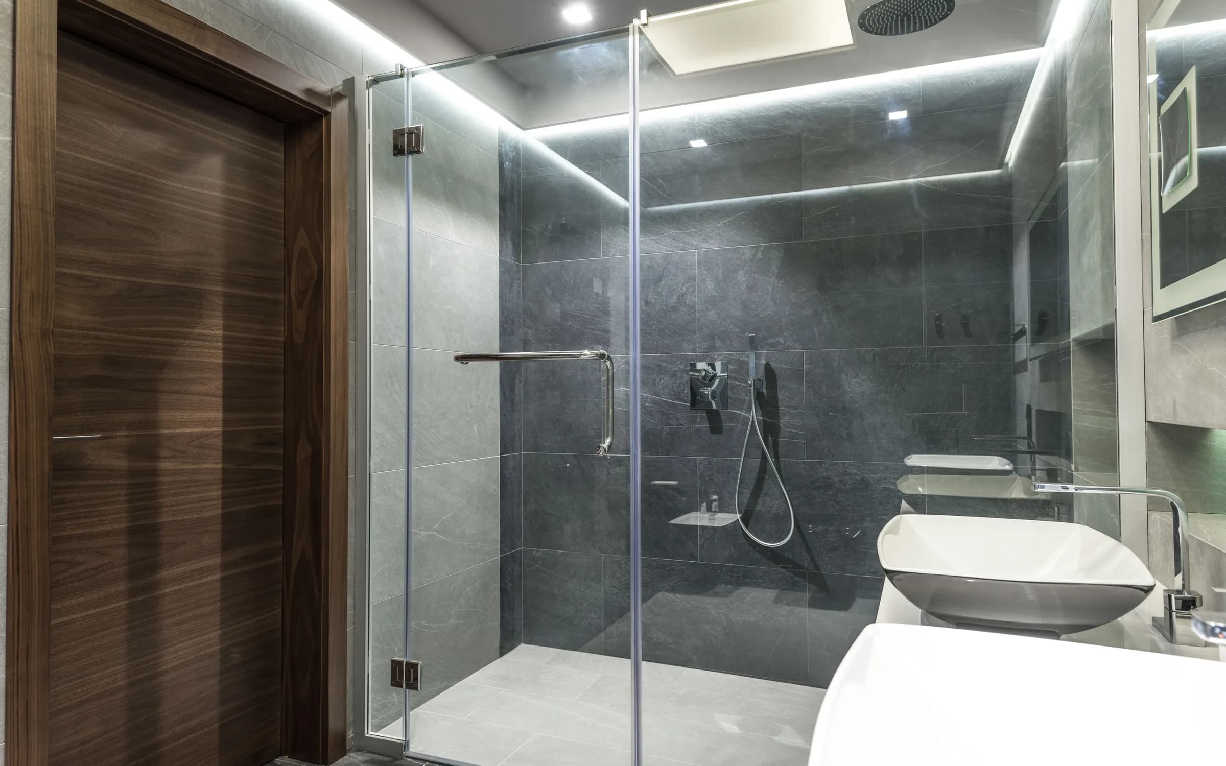 Koyu renk yüzeyleri, cam duşu ve çevresel aydınlatması bulunan modern banyo